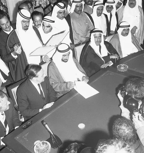 Men at a long table, one reading from a sheet, others crowding behind.