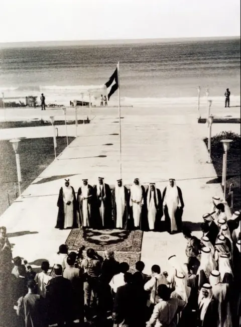 Seven Rulers standing on a carpet before the sea, a flag flying, photographers in front.